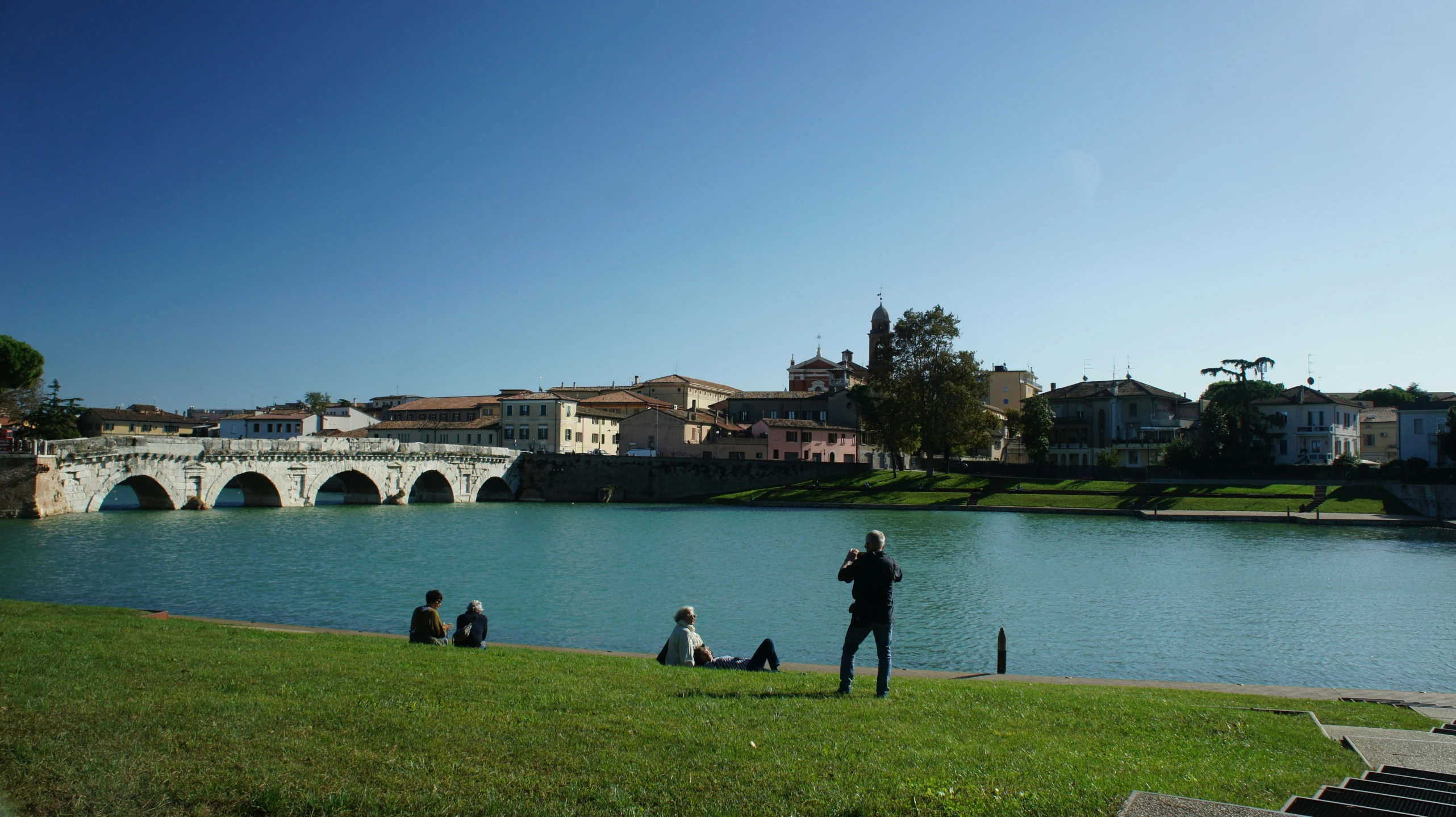 people sit and walk in the grass near a lake