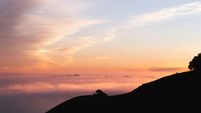 some clouds in the distance are below a lone tree on top of a mountain