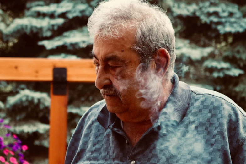 an old man smoking a cigarette in front of evergreens