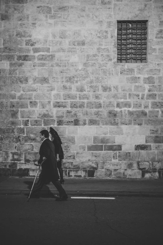 two people walking on the street next to a stone building