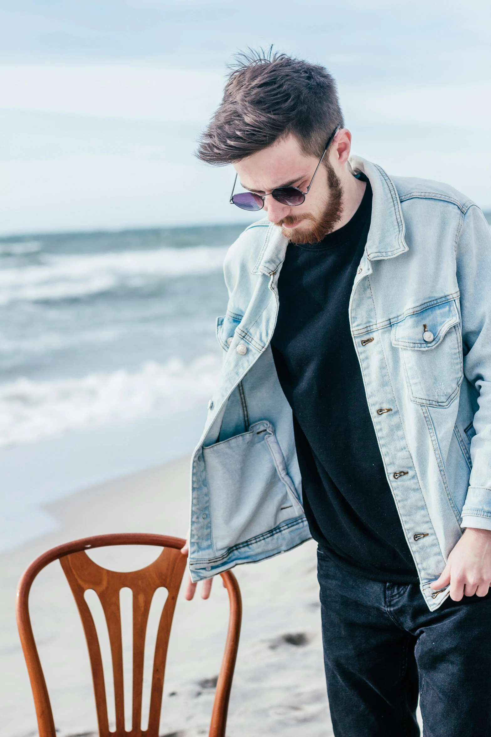 a man with beard leaning against a wooden chair at the beach