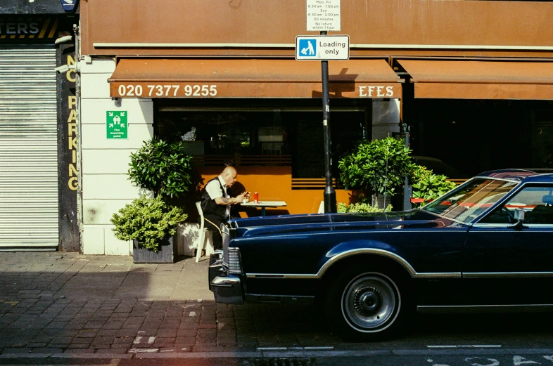 an older man sits on a chair in front of his car