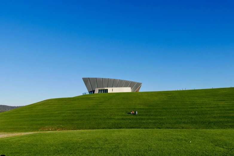 two people walking through the green grass on top of a hill