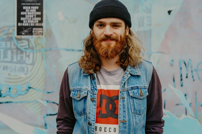 a young man with long hair and a beard wearing a denim vest