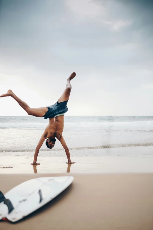 the man is doing a handstand on his feet on the beach near a surfboard