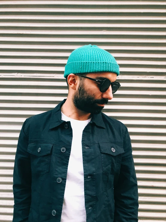 a bearded man wearing sunglasses, a black coat and a green beanie