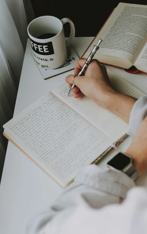 someone writing on notebooks at a table with a cup