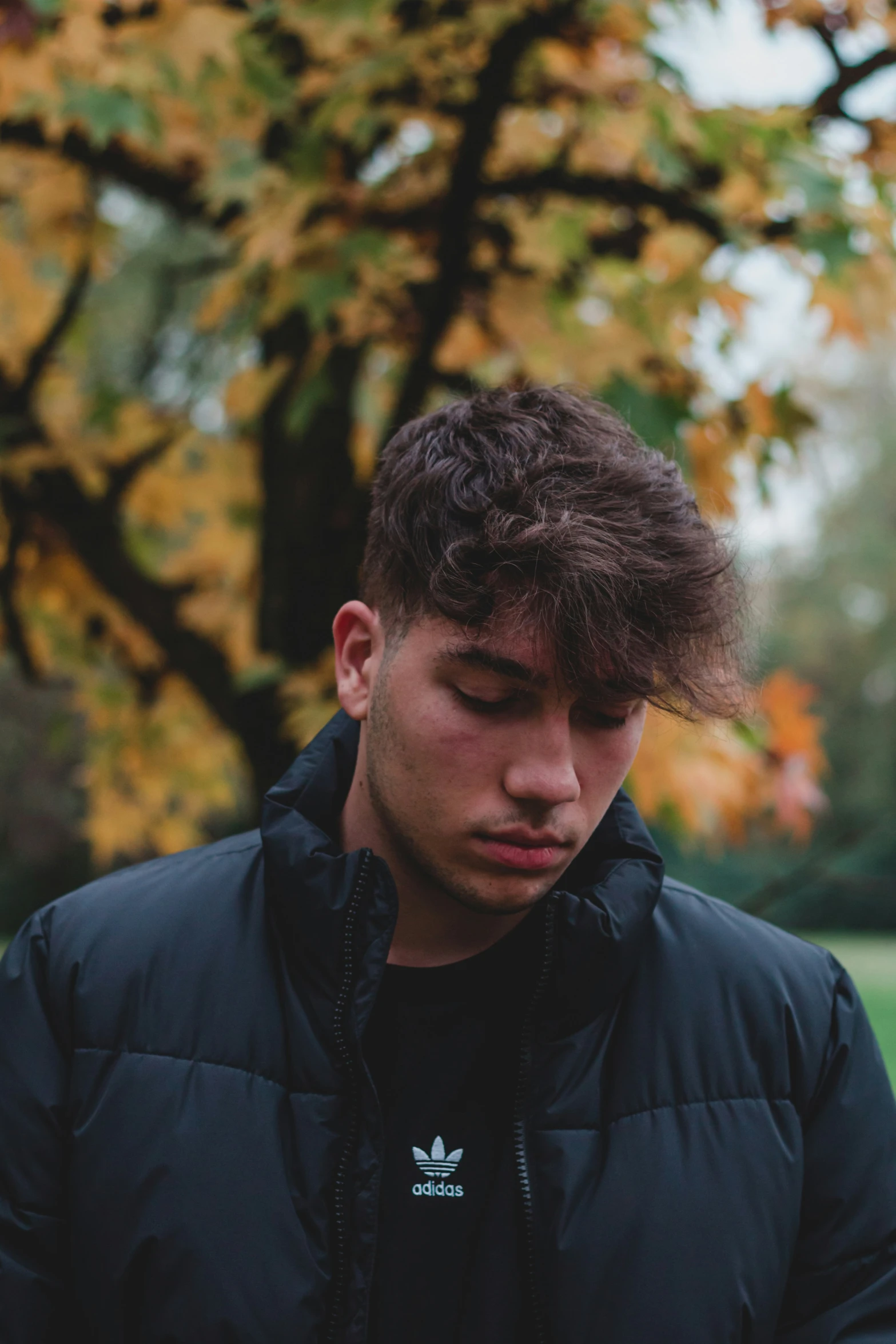 a man with a black jacket and blue jeans stands by the tree