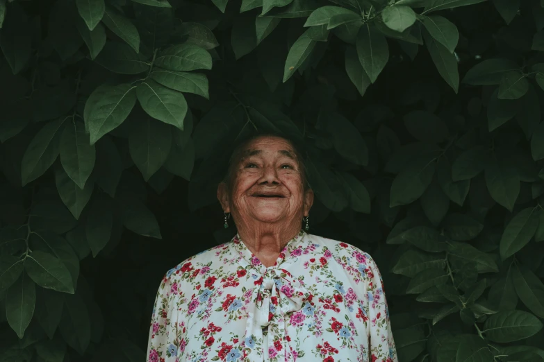 a smiling older woman standing by green leaves