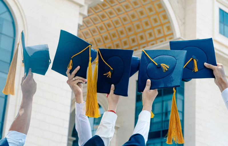 six graduating students hold up their caps in front of an ornately decorated building