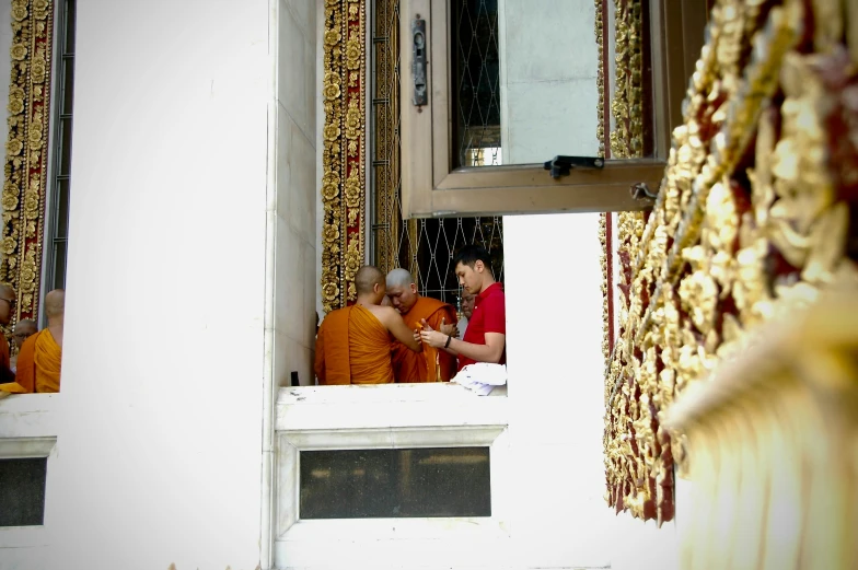 three monks in orange robes stand on a window sill, while a d over the window shows gold beads hanging from the sides of windows