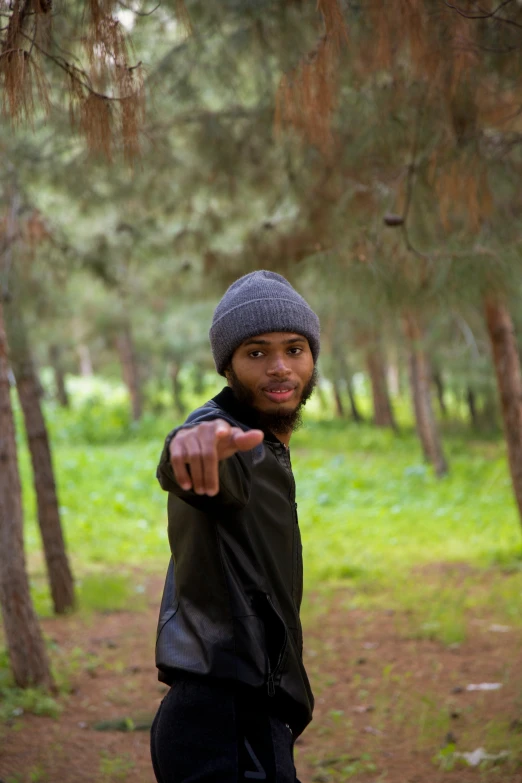 young man wearing a black jacket and a gray knit hat making a gesture in the woods