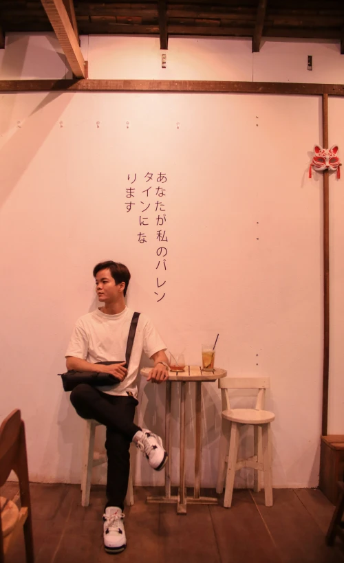 a man sits on a small wooden table next to a wall and some chairs