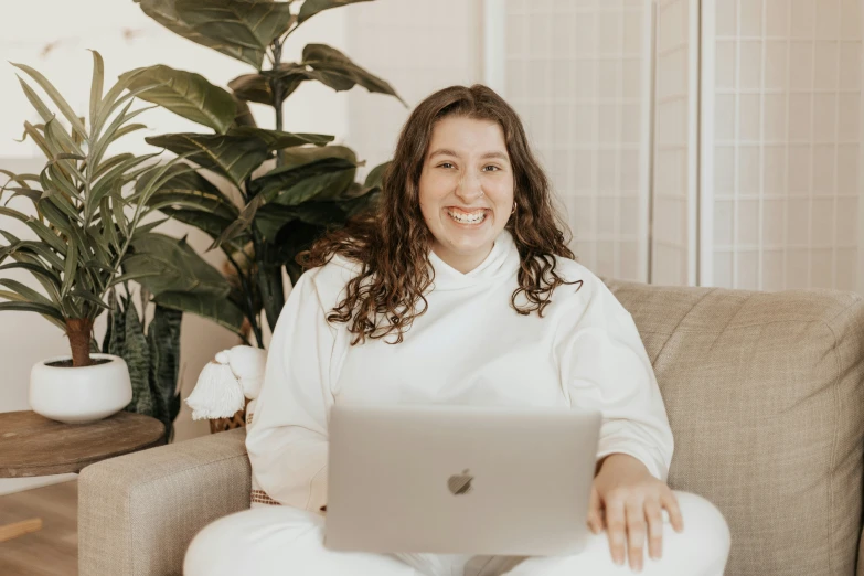 a young woman in white shirt and white pants smiling on couch with a laptop