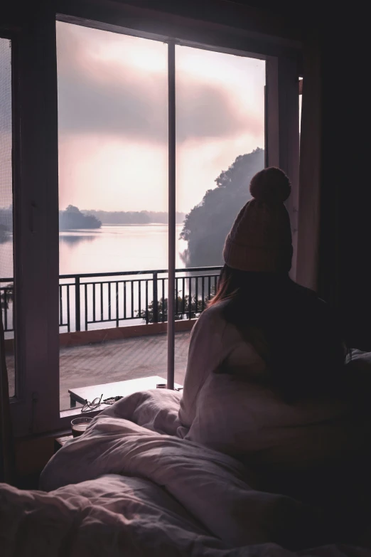 a woman sitting on her bed looking out onto a body of water