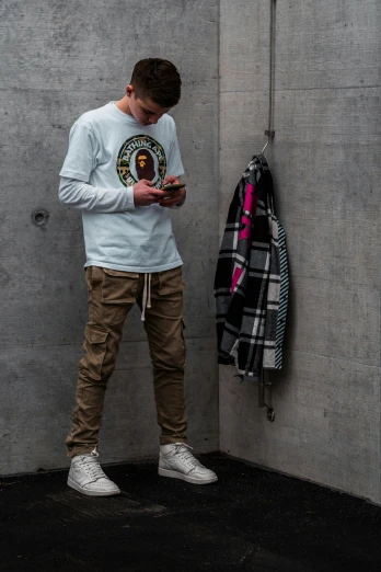 a boy in a white shirt and tan pants stands against a grey wall looking at his phone
