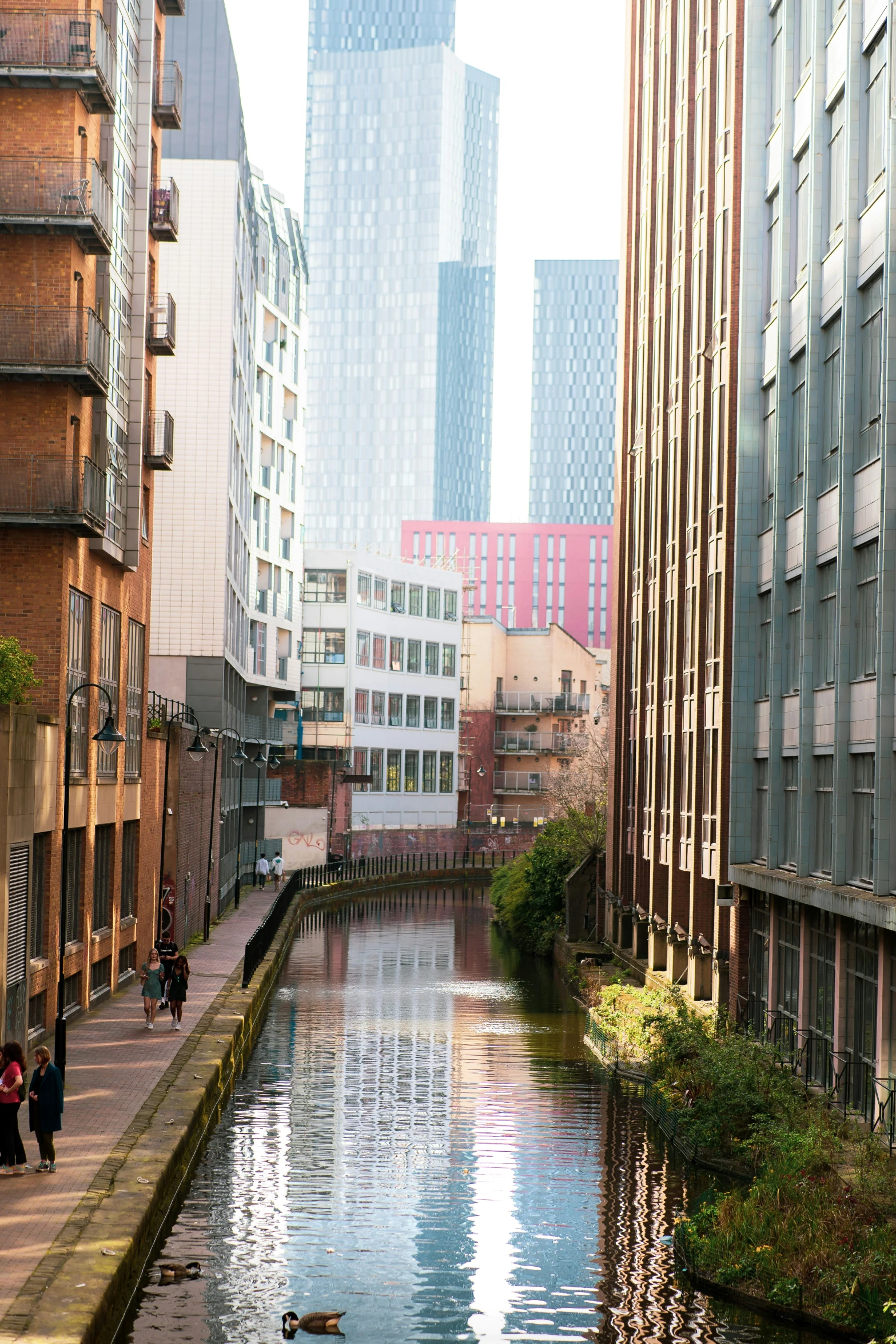 people walk around the area on one side of the waterway as tall buildings stand behind them