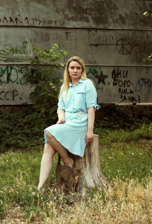 a blond woman in light blue dress posing next to wooden stump