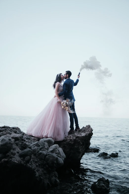 a couple stands on rocks holding smoke sticks while standing together