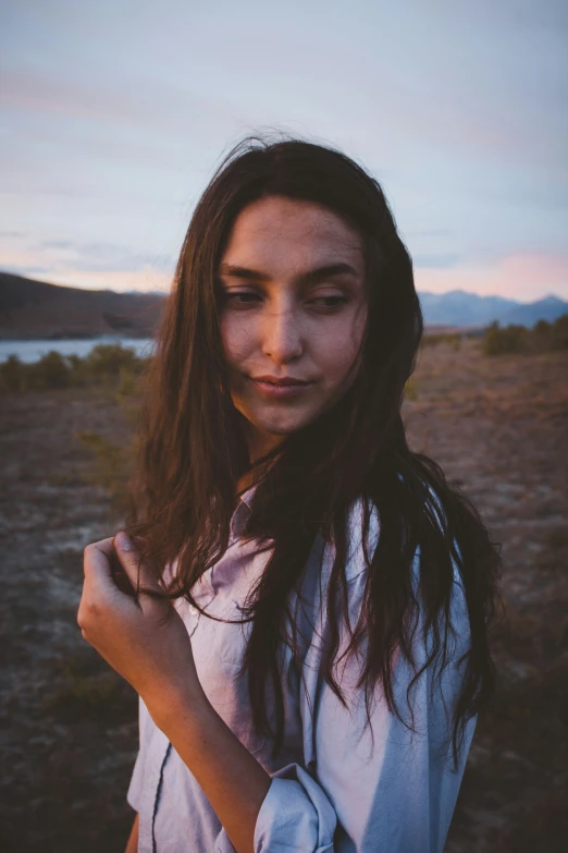 a woman in a white shirt and blue shirt with long hair stands in the middle of a dry field