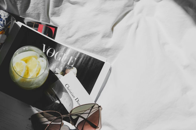 a book laying on top of a bed next to sunglasses and a drink