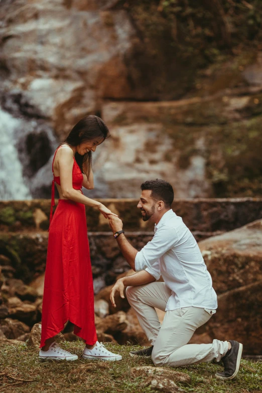 a woman in a red dress and a man in a white shirt hold hands at the bottom of a waterfall