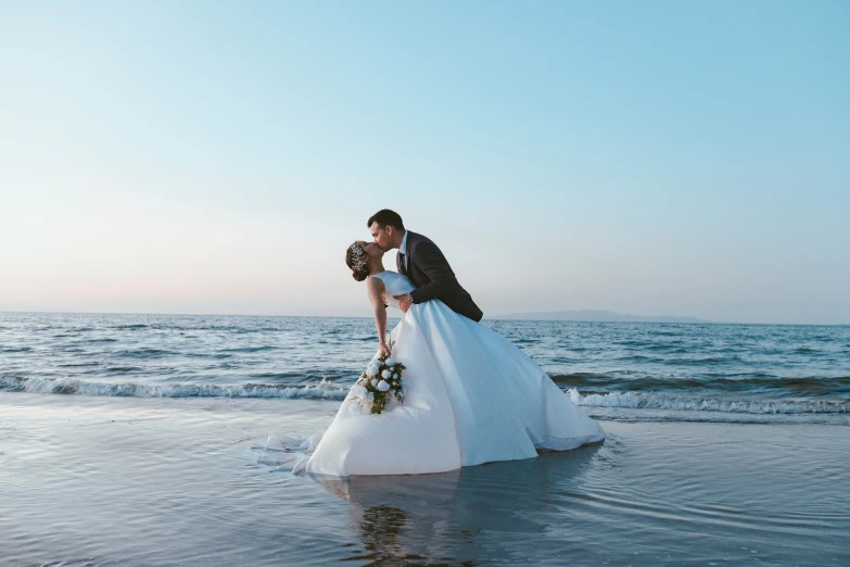 a bride and groom standing together on the shore of the beach