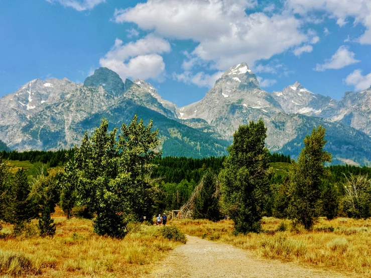 two hikers walk down a dirt path with mountains in the background