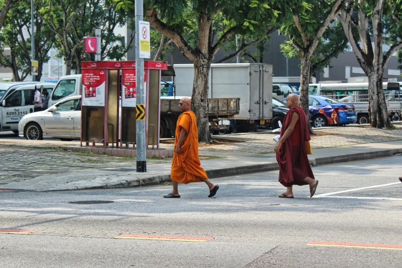 monks walking across the street with parked cars and buses behind them