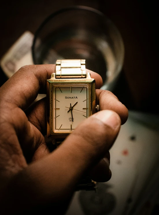 a hand holding a gold watch with a blurred glass in the background