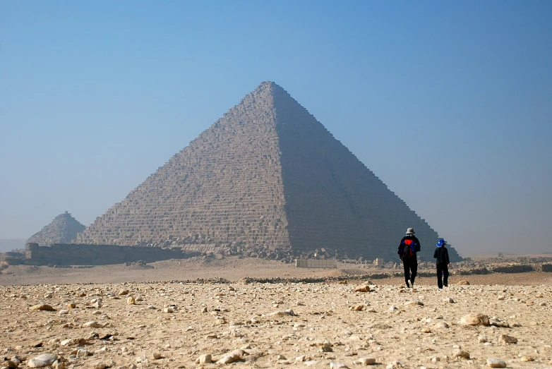 two people walking on a sandy area in front of a pyramid