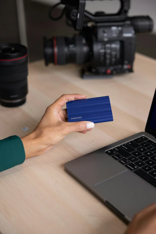 a hand holds a blue paper in front of a laptop