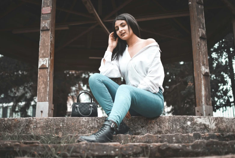 young woman in jeans, white blouse and black boots sitting on stairs talking on phone