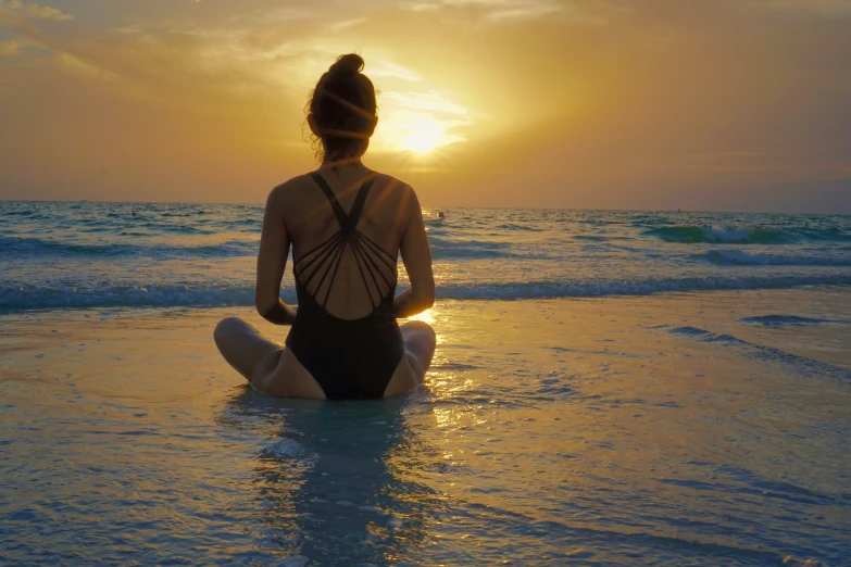 a woman is meditating on the beach during a sunset