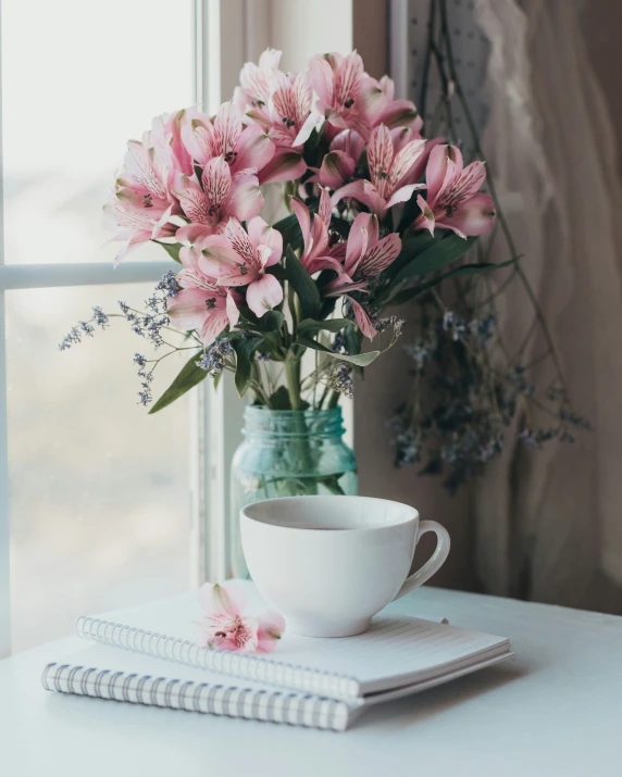 a bouquet of flowers sits on a notebook next to a glass vase and coffee mug