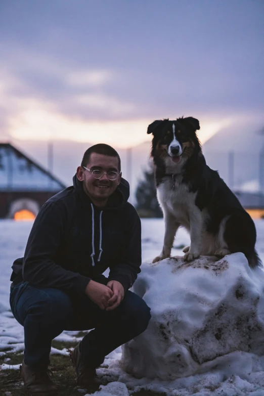 the man squats down next to his dog that stands in the snow