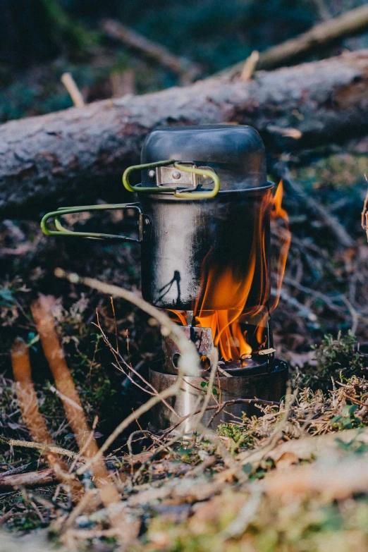 a camping stove with yellow flames and black cooking pot on the ground