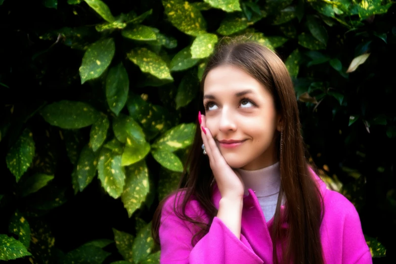 a girl wearing a pink top leaning up against a wall and looking into the distance with her hands to her face
