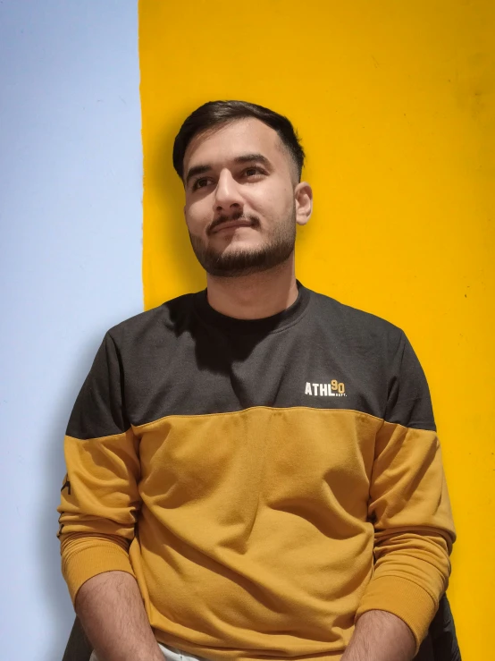 a man with a beard and mustard shirt on sitting against a wall