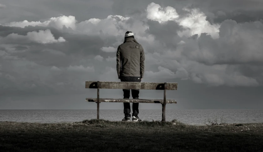 a man in an oversize coat looks out into the sea while sitting on a bench