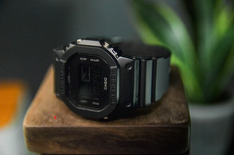 a digital watch sits on a wood block with a potted plant behind it