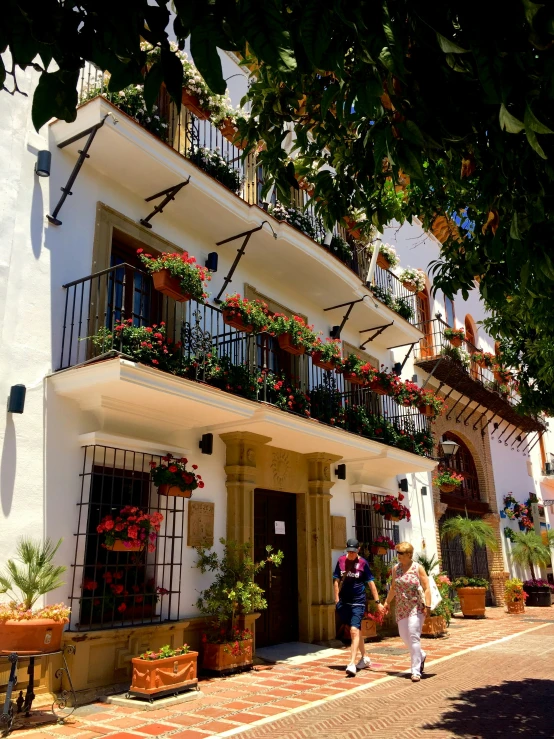 a couple of people walking past a building with plants on the balconies