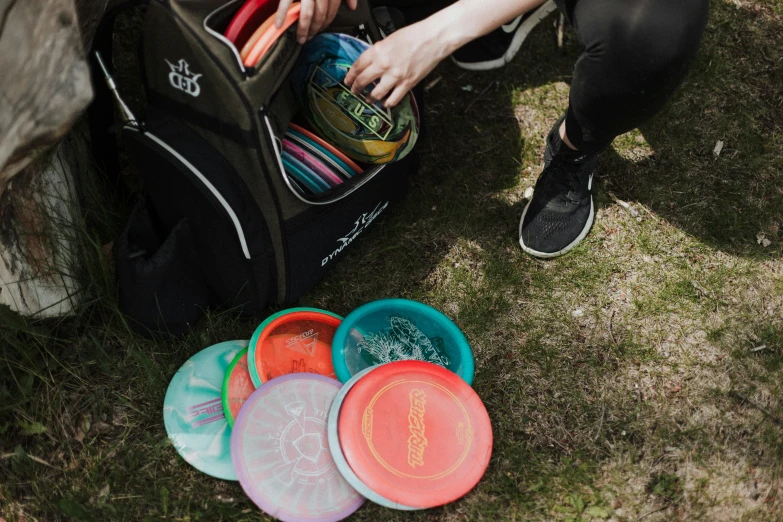 a man sits in the grass holding a duffel bag, with several frisbees on it