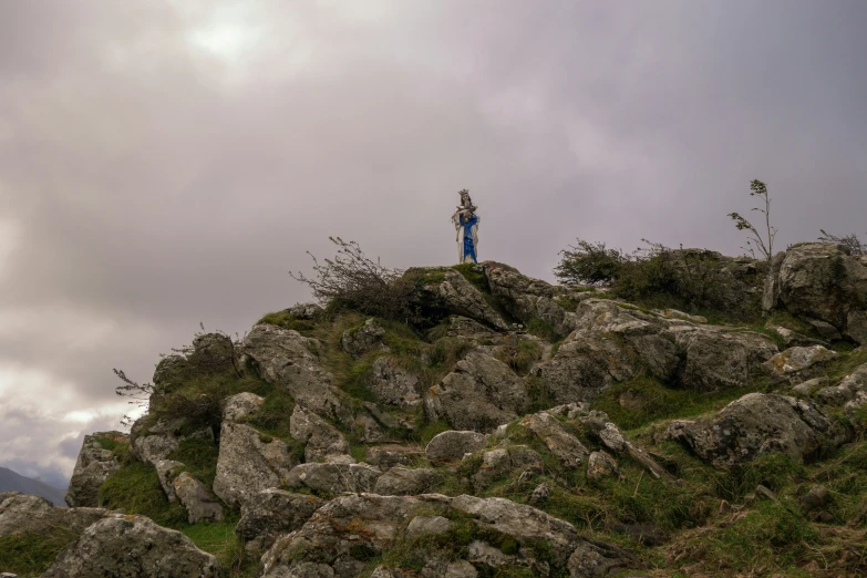 a person stands on the side of a rocky hill, surrounded by gray and green grass