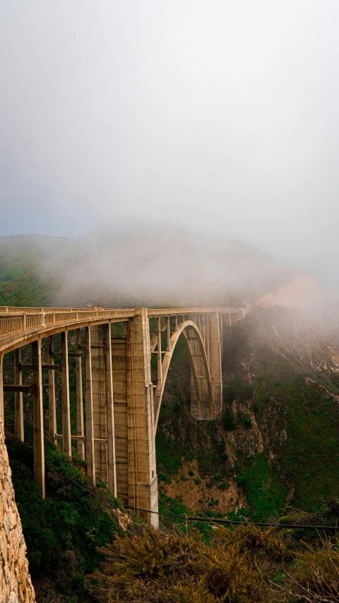 an arched bridge going over a valley in a foggy, misty day