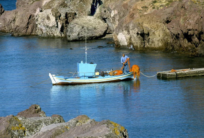 a man on a blue boat in the ocean near large rocks