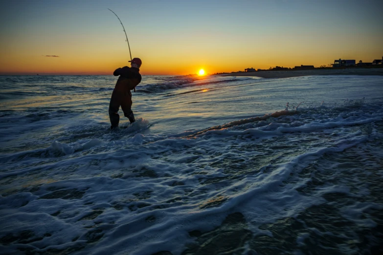 a man with fishing rod walks out of the ocean while sunsets