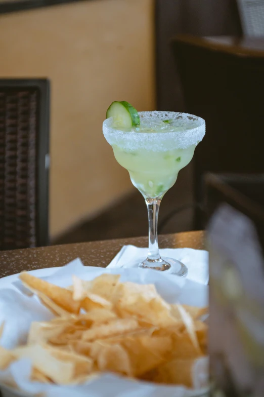 an alcoholic drink with cucumber garnish sits in a tall stemmed glass next to some chips