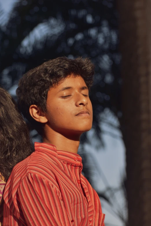 a young man wearing an orange shirt and standing next to a tree