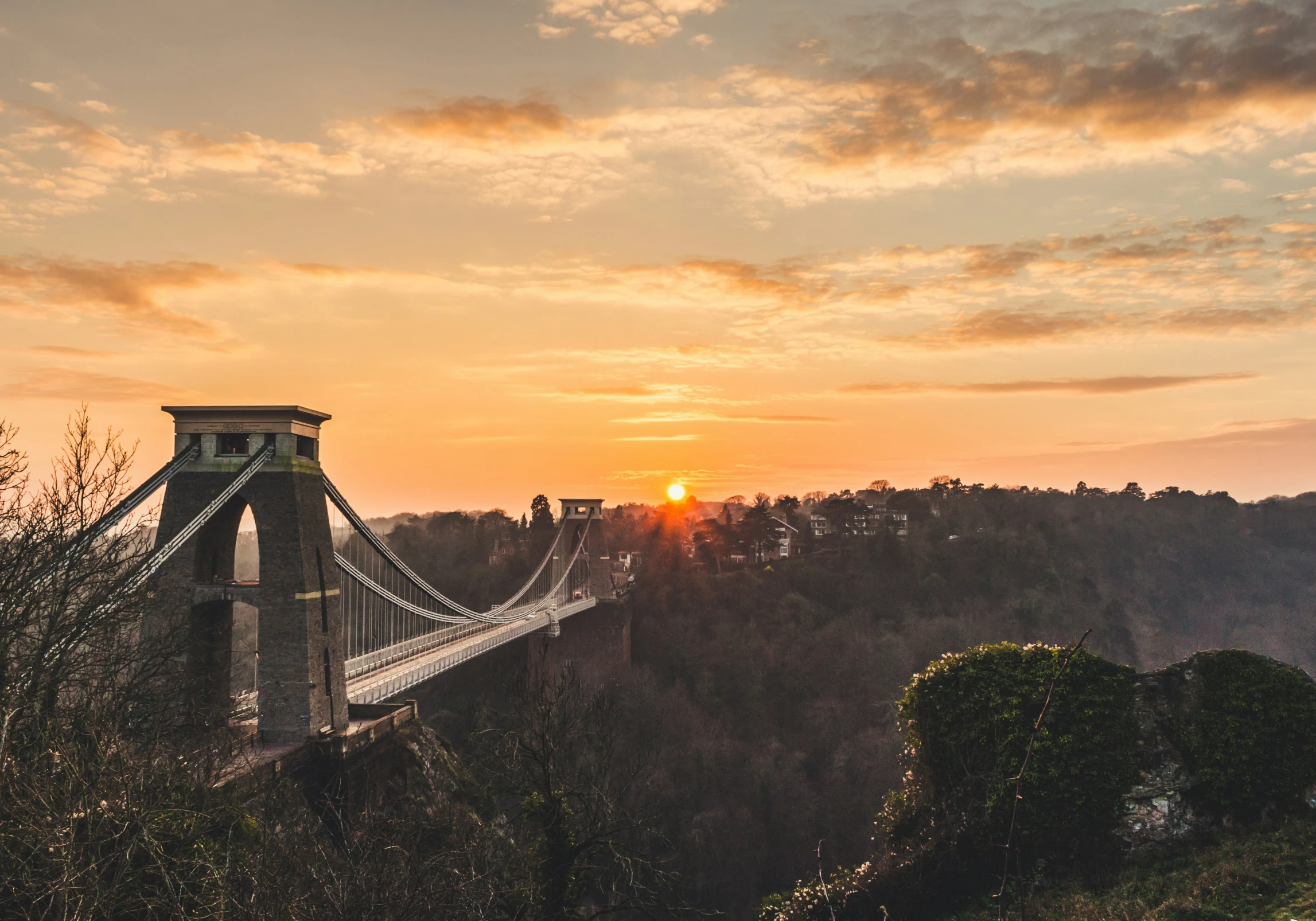 the sun rises over a tree covered hills and the bridge is over water